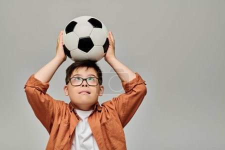Foto de Un niño encantador con síndrome de Down sostiene alegremente una pelota de fútbol sobre su cabeza. - Imagen libre de derechos