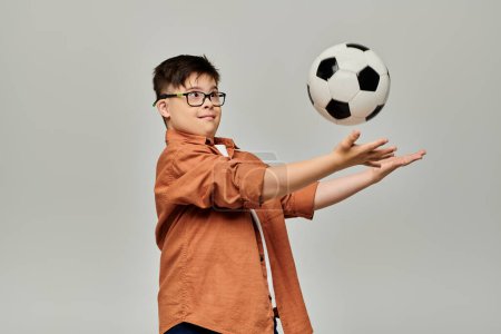 Foto de Un niño encantador con síndrome de Down equilibra una pelota de fútbol sobre un fondo gris. - Imagen libre de derechos