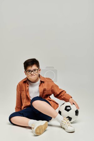 Foto de Little boy with with Down syndrome with glasses sits on ground with soccer ball. - Imagen libre de derechos