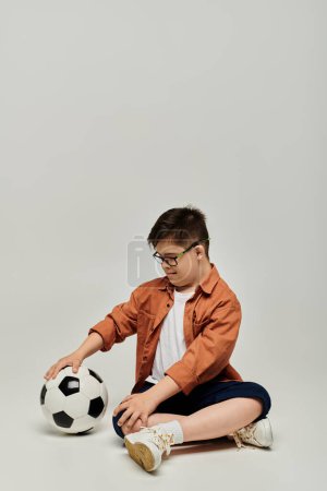 Téléchargez les photos : Petit garçon avec le syndrome de Down assis sur le sol avec un ballon de football. - en image libre de droit