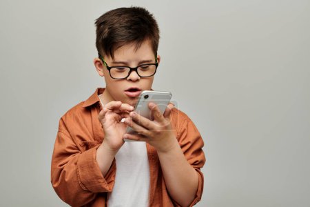 Foto de Little boy with Down syndrome wearing glasses focuses intently on smartphone screen. - Imagen libre de derechos