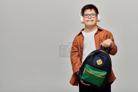 Ein kleiner Junge mit Down-Syndrom hält einen Rucksack und trägt Kopfhörer.