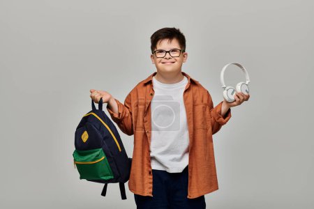 Un niño pequeño con síndrome de Down sosteniendo una mochila y usando auriculares.