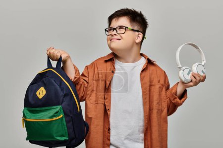 little boy with Down syndrome holds backpack and headphones.