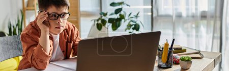 Téléchargez les photos : Un garçon concentré avec le syndrome de Down est assis à un bureau, regardant un ordinateur portable devant lui. - en image libre de droit