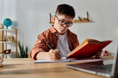 A boy with Down syndrome sits at a desk with a laptop and a book.