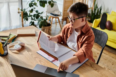 Foto de Adorable chico con síndrome de Down absorto en el ordenador portátil y libro en la mesa. - Imagen libre de derechos