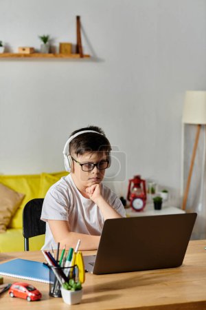 A boy with Down syndrome sitting at a table, wearing headphones and using a laptop.