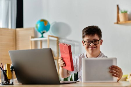 Téléchargez les photos : Un garçon trisomique assis à un bureau avec un ordinateur portable et un livre. - en image libre de droit