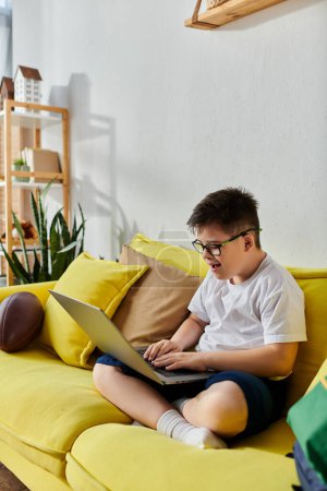 Foto de Un niño con síndrome de Down usa una computadora portátil en un sofá amarillo brillante. - Imagen libre de derechos