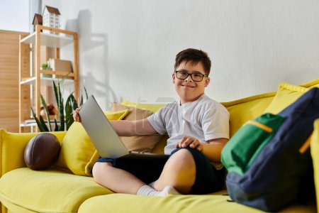 Ein entzückender Junge mit Down-Syndrom sitzt auf einer gelben Couch und benutzt einen Laptop.