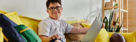 Photo for Adorable boy with Down syndrome sitting on yellow couch, using laptop. - Royalty Free Image