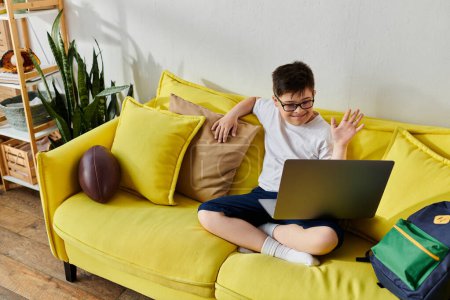 adorable chico con síndrome de Down utiliza portátil en sofá amarillo en casa.