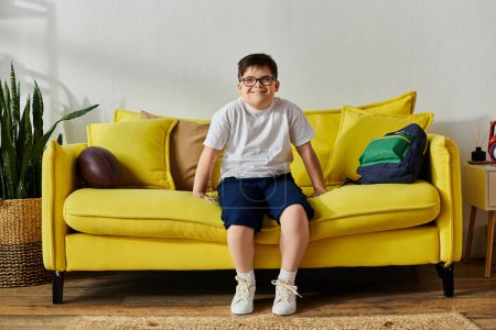 Foto de Un chico lindo con síndrome de Down sentado en un sofá amarillo brillante en casa. - Imagen libre de derechos