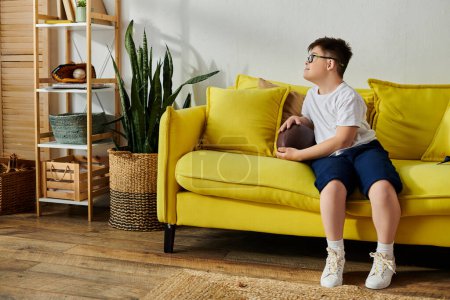 A adorable boy with Down syndrome happily sits on a vibrant yellow couch in his room.