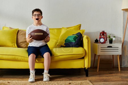 Ein entzückender Junge mit Down-Syndrom hält einen Fußball in der Hand, sitzt auf einer leuchtend gelben Couch.