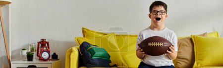 Foto de Little boy happily holds a football while sitting on a couch at home. - Imagen libre de derechos