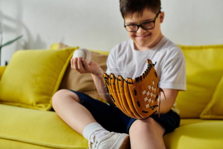 Foto de Un niño con síndrome de Down se sienta en un sofá con un guante de béisbol. - Imagen libre de derechos