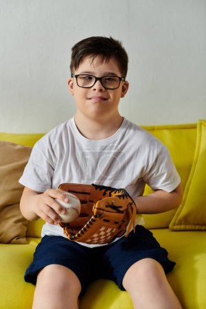 Téléchargez les photos : Adorable garçon avec trisomie 21 avec des lunettes assis sur un canapé jaune tenant une balle de baseball. - en image libre de droit