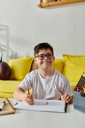 adorable boy with Down syndrome in glasses, surrounded by pens and pencils, sits at table engrossed in drawing or writing.