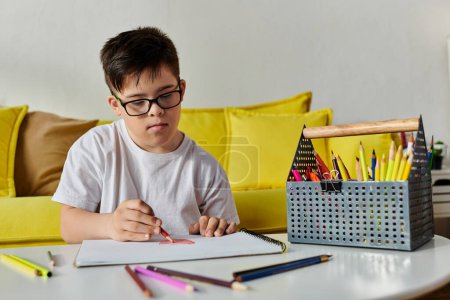 Un garçon trisomique dans des lunettes dessinant avec des crayons de couleur à une table dans sa chambre.