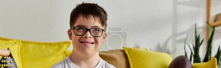 Foto de Un niño adorable con síndrome de Down con gafas se sienta tranquilamente en un sofá en una habitación acogedora. - Imagen libre de derechos