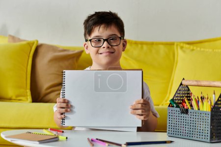 A adorable boy with Down syndrome proudly displays a sheet of paper in front of a yellow couch.