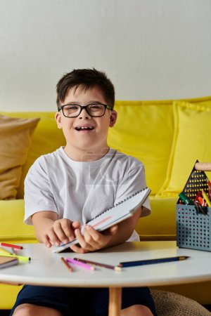 entzückender Junge mit Down-Syndrom mit Brille am Tisch, Färbung im Notizbuch mit Buntstiften.