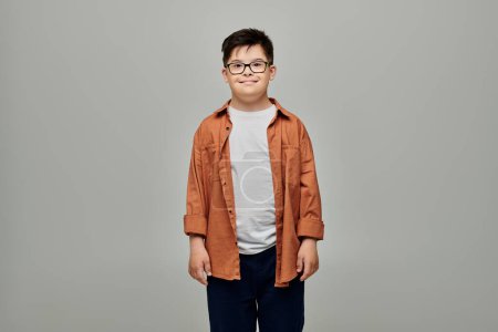 A charming little boy with Down syndrome wearing glasses stands against a gray background.