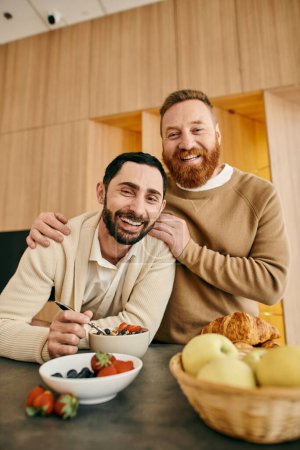 Two men strike a pose in front of a colorful bowl of fresh fruit, exuding joy and connection in a modern setting.