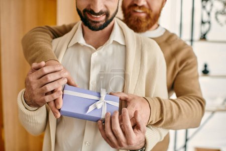 Two men embracing each other while tenderly holding a gift box in a modern apartment, embodying love and happiness.