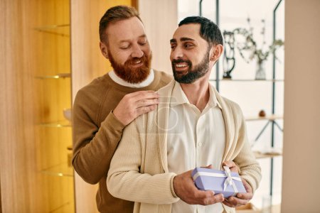 Dos hombres abrazan, sosteniendo una caja de regalo, expresando amor y aprecio en un ambiente de apartamento moderno.