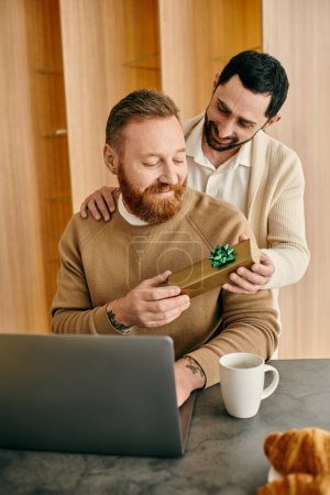 Two men exchange a gift in front of a laptop in a modern apartment, sharing a moment of happiness and love.