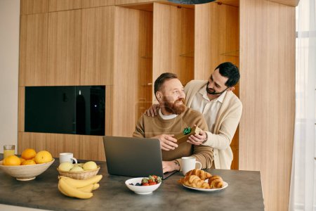 Zwei Männer arbeiten fröhlich an einem Laptop in der schlichten Küche einer modernen Wohnung.