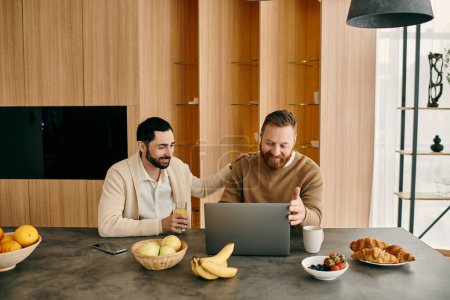 Photo for Two happy men, a gay couple, sit at a table in a modern apartment, engrossed in using a laptop. - Royalty Free Image