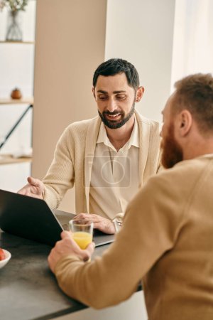 Photo for A happy gay couple sits at a table in a modern apartment, focusing on their laptop - Royalty Free Image