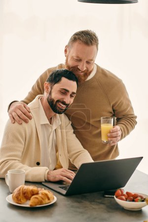 Zwei Männer, ein glückliches schwules Paar, sitzen an einem Tisch mit einem Laptop und arbeiten. Sie genießen auch Gläser Orangensaft.