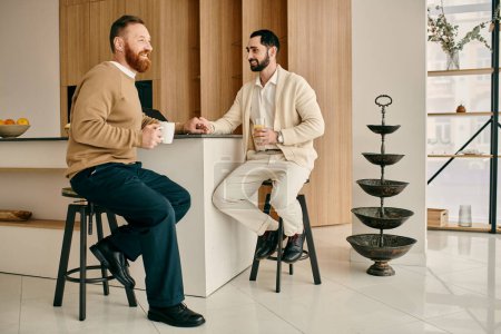 Dos hombres, una feliz pareja gay, conversan sentados en taburetes en una cocina moderna.