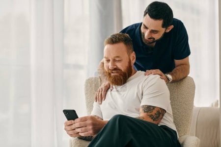 Foto de Dos hombres con ropa casual se sientan en una silla, uniéndose mientras miran un teléfono celular juntos en una sala de estar moderna. - Imagen libre de derechos