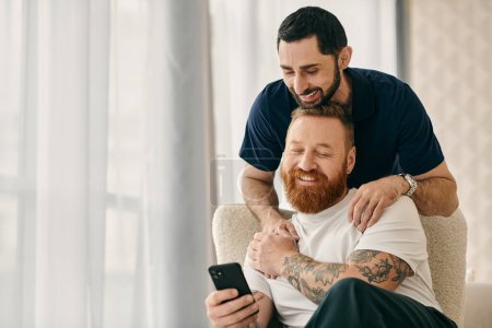 Foto de Two men in casual clothes sit on a couch, engrossed in a cell phone together in a modern living room. - Imagen libre de derechos