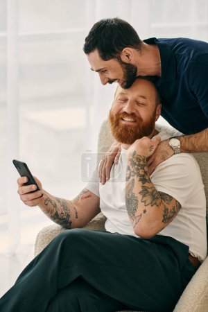 Foto de Two happy men with beards sit closely together on a chair in a modern living room, showing their love and affection. - Imagen libre de derechos
