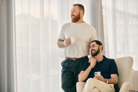 A happy gay couple in casual clothes sitting on a chair, drinking coffee, and sharing a moment in a modern living room.