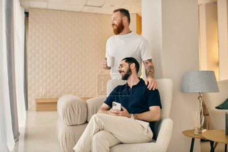 Foto de Dos hombres, una feliz pareja gay, en ropa casual se sientan juntos en una silla en una sala de estar moderna, apreciando el tiempo de calidad pasado. - Imagen libre de derechos