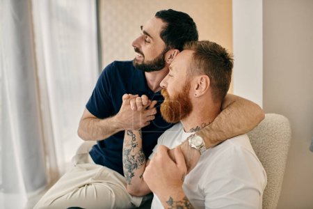 Foto de Two happy men with tattoos on their arms sit close on a couch in a modern living room, enjoying quality time together. - Imagen libre de derechos