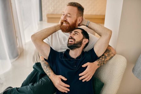 Foto de Una alegre pareja gay, en atuendo casual, compartiendo un cálido abrazo en un moderno sofá de la sala de estar. - Imagen libre de derechos