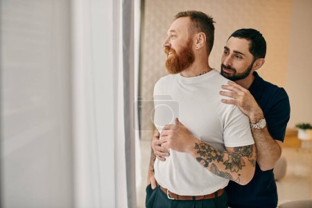 Foto de Two men in casual clothing embrace in a warm hug in front of a window, showcasing their love in a modern living room setting. - Imagen libre de derechos
