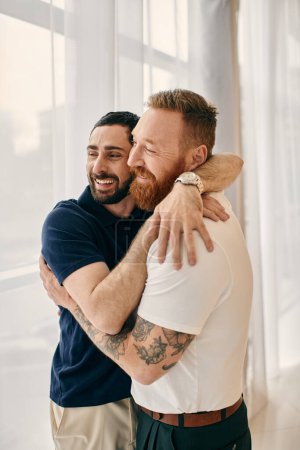Two men in casual clothes embrace each other warmly in front of a window in a modern living room.