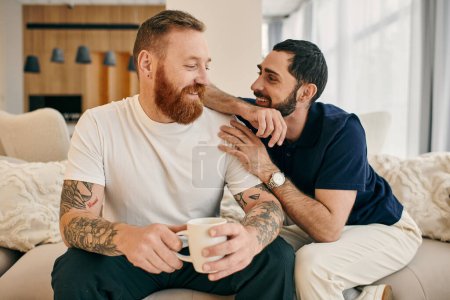 Foto de Una pareja gay en atuendo casual relajarse en un sofá, disfrutar de la compañía de los demás y tomar un café en un ambiente moderno sala de estar. - Imagen libre de derechos