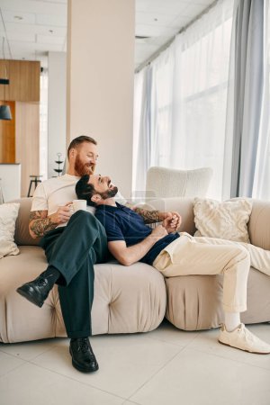Foto de Dos hombres, una feliz pareja gay, vestidos casualmente, se sientan juntos en un moderno sofá de la sala de estar, disfrutando de la compañía de los demás. - Imagen libre de derechos