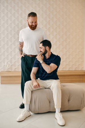 Foto de Dos hombres, una feliz pareja gay, se sientan uno al lado del otro en una moderna sala de estar, compartiendo tiempo de calidad juntos. - Imagen libre de derechos